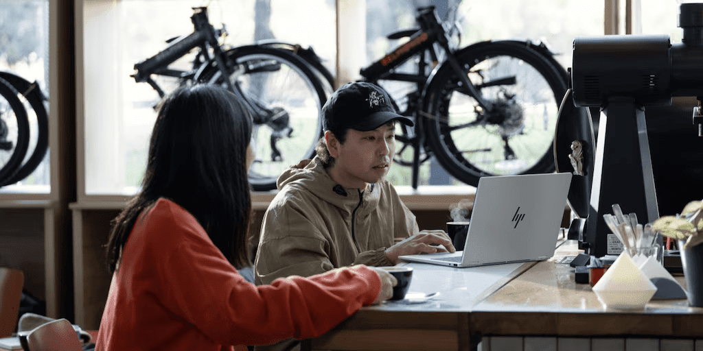 Two people working together at a counter with an HP laptop in a bicycle shop, with bikes hanging on display in the background