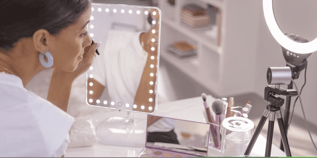 A woman applying makeup while looking into an illuminated vanity mirror with a webcam and ring light setup for beauty content creation.RetryClaude can make mistakes. Please double-check responses.